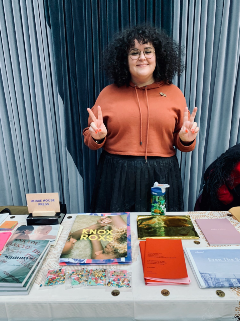 Photograph of Adriana Monsalve behind a table covered with books. The artist is smiling and giving a double peace sign.