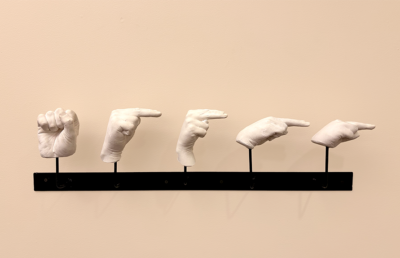 Five sculptures of hands, cast in white plaster, spell out the ASL letters for "SHHHH". They are mounted on black brackets against an off-white wall.